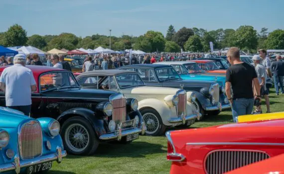 A group of classic cars displayed outdoors with people walking around and admiring them on a sunny day at a motor show.