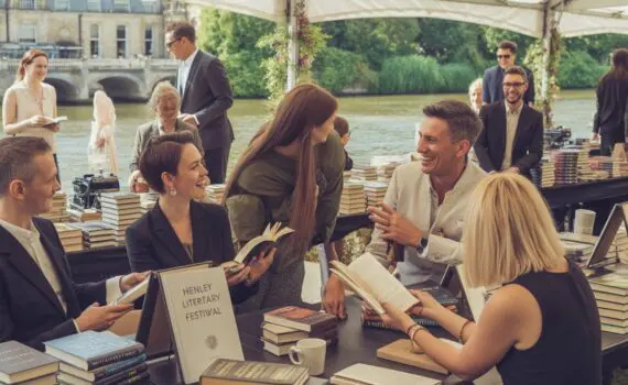 People enjoying a literary festival outdoors by a river with tents, books, and greenery in the background.